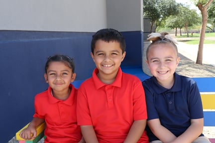 Three kids sit together on a colorful bench outdoors, smiling at the camera.