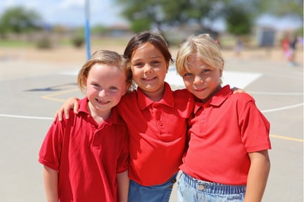Three young students wearing red shirts stand close together outdoors, smiling with their arms around each other on a sunny school playground.